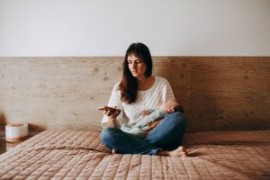 A mother sits on a bed holding her baby in a serene bedroom setting, checking her phone.